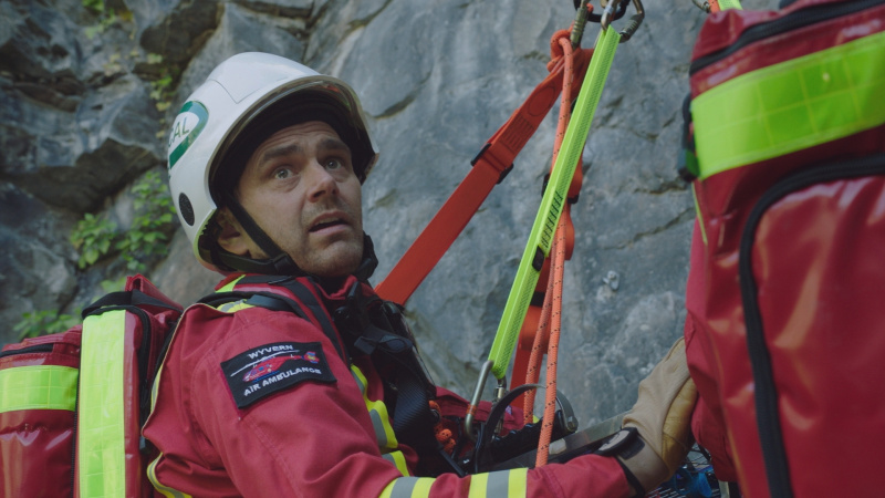 Iain Dean îmbrăcat în uniformă roșie HEMS pe o stâncă în episodul Casualty Precipice.