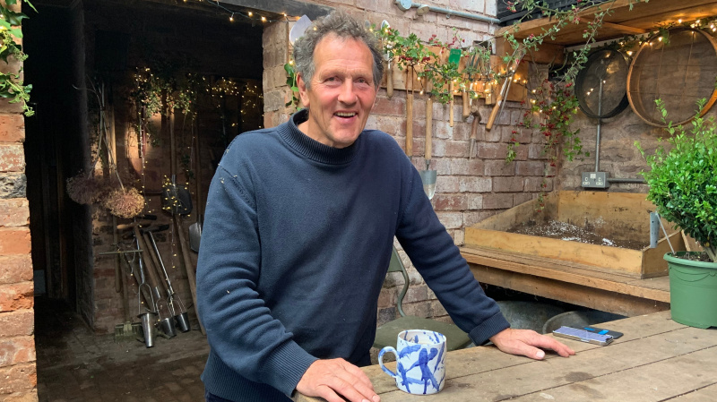   Monty Don trägt einen dunkelblauen Pullover und trinkt eine Tasse Tee im Garten