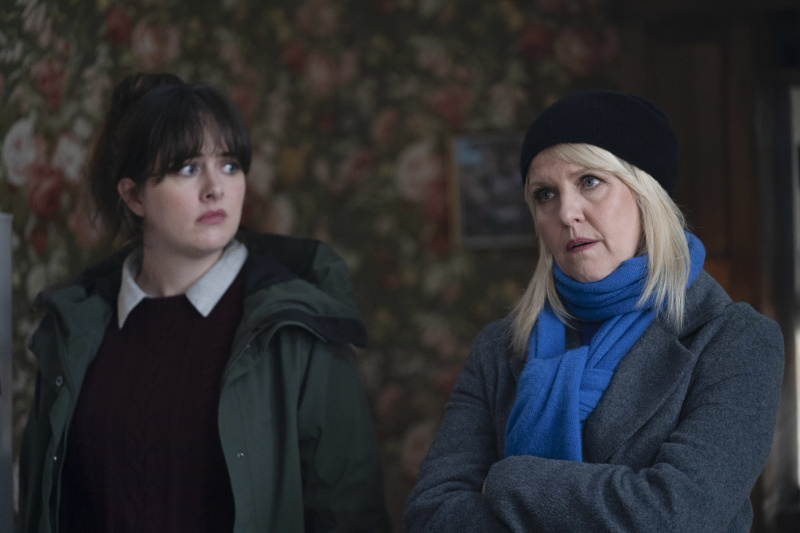   Tosh (Alison O'Donnell) and Calder (Ashley Jensen) stand inside a house, both still wearing their outdoor coats, with Calder also wearing a scarf and wool hat, while Tosh's coat is unfastened. Calder has her arms folded as she speaks to an unseen suspect, while Tosh is looking at Calder