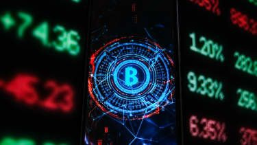 Ένα λογότυπο Bitcoin εμφανίζεται σε ένα smartphone με τα ποσοστά του χρηματιστηρίου στο φόντο
