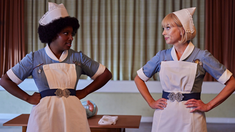   Joyce a Trixie bok po boku v uniformách pôrodnej asistentky, keď sa na seba pozerajú s rukami v bok v 14. sezóne Call the Midwife