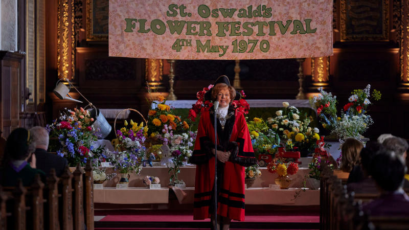   Fialová v červenom starostovskom rúchu stojaca pred kostolom a otvárajúca kvetinový festival v sezóne Call the Midwife 14 ep 2