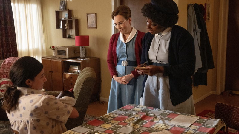   Üniformalı Shelagh (Laura Main) ve Joyce (Renee Bailey), Ebeyi Ara'da bebeğini kucağına alan bir anneyle konuşuyor.