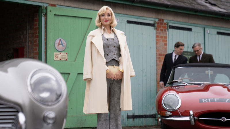   Helen George ในเสื้อคลุมสีขาวและชุดสูทสีเทายืนอยู่ระหว่างรถสองคันในเรื่อง Call the Midwife