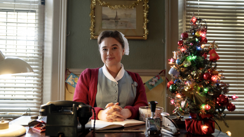   Megan Cusack ões's outfit as Nancy sits at the surgery reception desk beside a Christmas tree in the Call the Midwife Christmas Special 2024.
