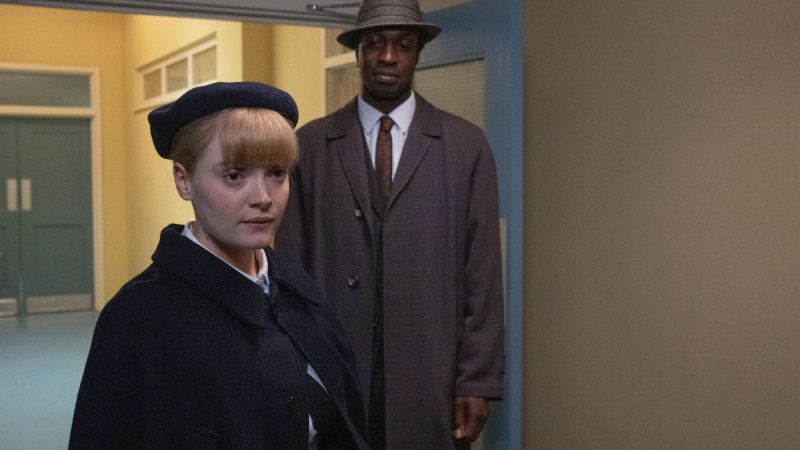   Rosalind (Natalie Quarry) v uniforme stojí v hale s Cyrilom (Zephryn Taitte) v tmavom obleku a klobúku vo filme Call the Midwife.