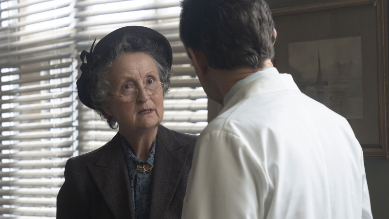   Georgie Glen v tmavom obleku, klobúku a okuliaroch ako slečna Higginsová sa rozpráva so Stephenom McGannom v bielom plášti ako Dr Turner vo filme Call the Midwife.
