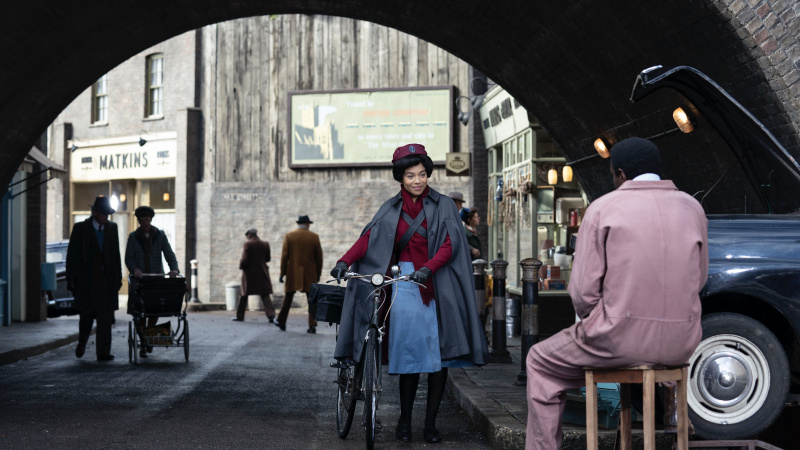   Leonie Elliott em enfermeira's uniform as Lucille pushes a bike towards Zephryn Taitte in overalls as Cyril in Call the Midwife.