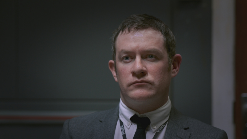   Philip Hill-Pearson als detective Bell die er serieus en streng uitziet in een grijs pak en grijze ondervragingsruimte.