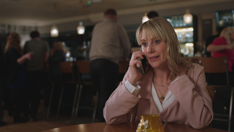   Kellie Shirley als verpleegster Sophia Peters die in een pub zit in een witte blouse en roze jas, aan de telefoon en er van streek uitzien.