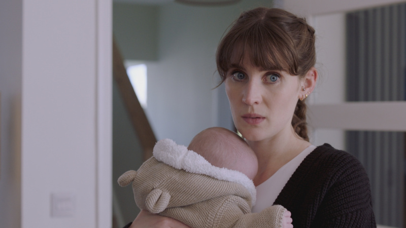   Anna Chell als Jodie Whyte, die een baby in een warme onesie houdt terwijl ze uit grote ogen ziet en bezorgd.