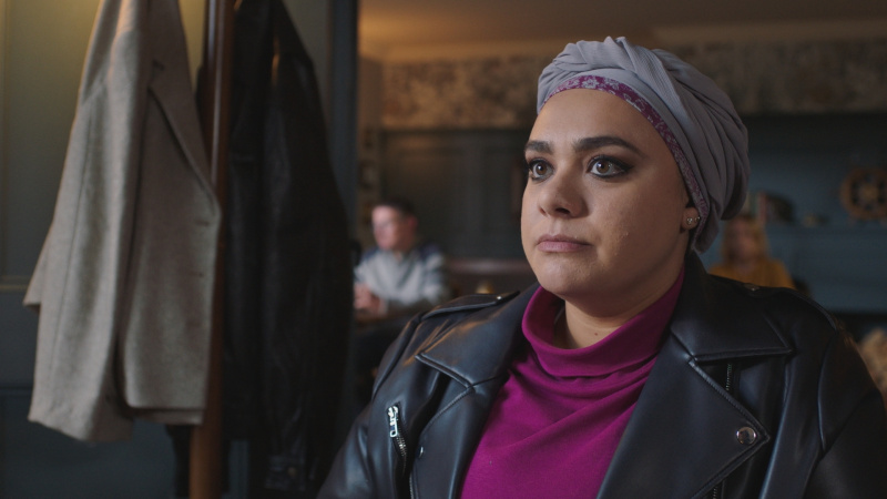   Sarah Seggari als verpleegster Rida Amaan in een pub die streng uitziet en een lila -hoofddoek, paarse polo nek en zwart jasje draagt.