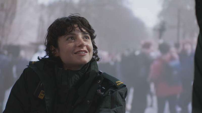   Naomi Wakszlak en tant qu'ambulancier paramédical stagiaire Indie Jankowski dans Casualty 2025, portant l'uniforme vert d'ambulancier complet et souriant à quelqu'un hors écran.