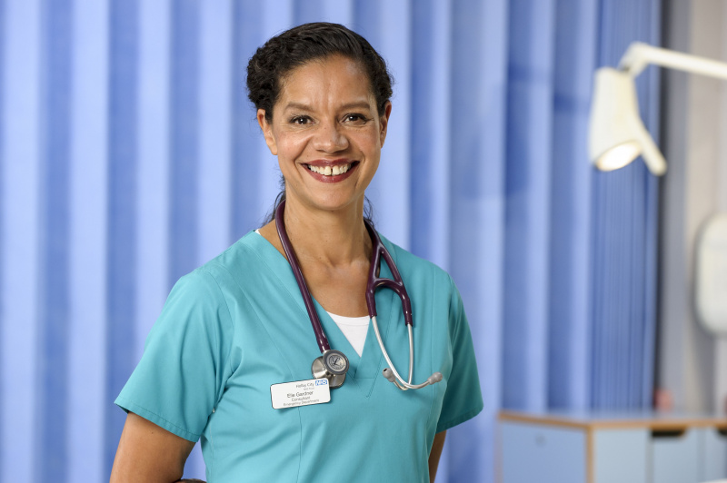  Jaye Griffiths souriante dans le rôle du docteur Elle Gardner dans Casualty, portant une blouse bleu vif.