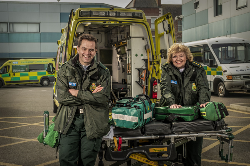   Poseerattu otos Michael Stevensonista ja Di Botcherista, jotka nojaavat avoimen ambulanssin selkään ensihoitajavarusteissa heidän Iain Deanina ja Jan Jenninginä Casualty-elokuvassa.