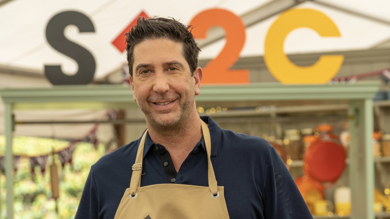   Дейвид Швимер в тъмен топ и бледа престилка в палатката за The Great Celebrity Bake Off за Stand Up to Cancer