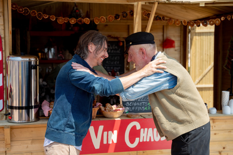   Christian (Philippe Spall) og Frederick (Pavel Douglas) står ved siden av en gløggbod på julemarkedet i Sainte Victoire, gliser og lener seg på hverandre's shoulders