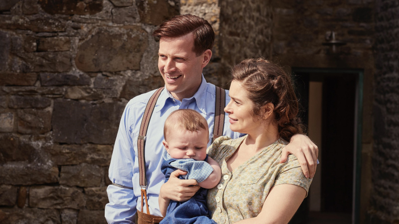 Nicholas Ralph v modrej košeli ako James stojí s Rachel Shentonovou v béžovej blúzke a drží malého Jimmyho vo filme All Creatures Great and Small.