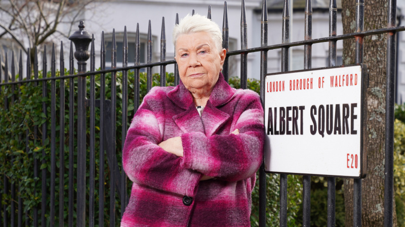   Mo Harris con le braccia incrociate in posa accanto all'insegna di Albert Square e indossa un cappotto a quadri rosa.