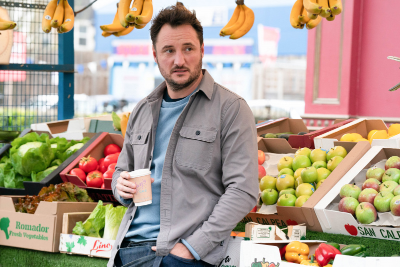  Martin Fowler tomando café y apoyado en su puesto de frutas y verduras.