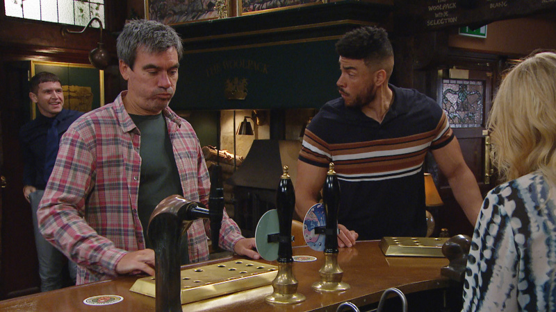   Nate Robinson și Cain Dingle la barul The Woolpack. Corey zâmbește în spatele lor în timp ce Charity Dingle scoate o halbă.