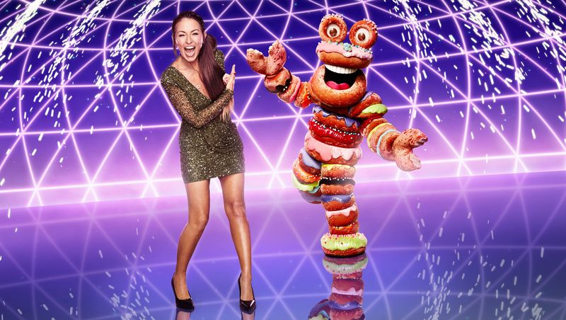 Davina McCall stovi priešais purpurinį foną šalia Kaukės dainininkės personažo Donuts – kostiumo, sudaryto iš spurgų žiedų, sukrautų viena ant kitos.