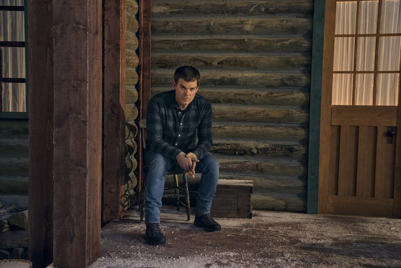Michael C. Hall ruudullisessa paidassa ja farkuissa istumassa mökin ulkopuolella