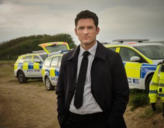 The Long Call - Ben Aldridge como DI Matthew Venn, em uma camisa e gravata com um longo casaco escuro, parado em uma praia de Devon com carros da polícia atrás dele