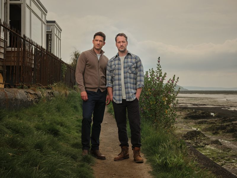 Matthew Venn (Ben Aldridge) com seu marido Jonathan (Declan Bennett) em pé no caminho do lado de fora de sua casa, com a praia ao fundo