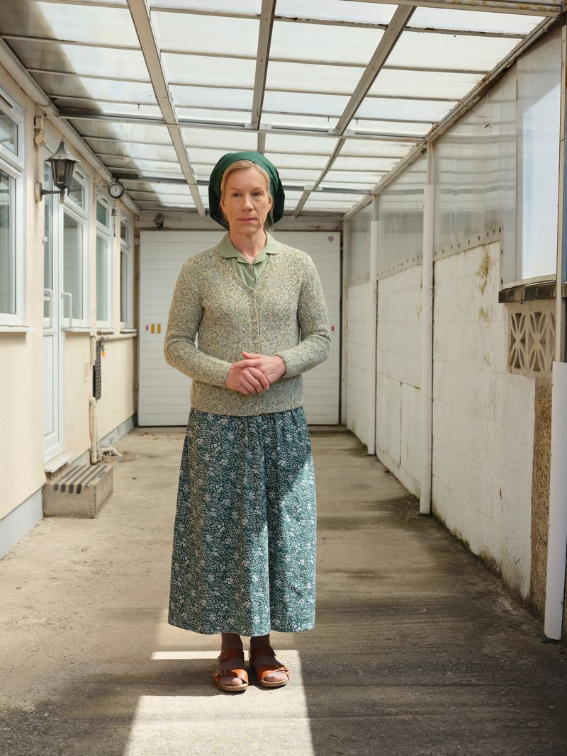 Juliet Stevenson como Dorothy Venn, vestindo um cardigã, com uma saia longa e um lenço na cabeça, do lado de fora de um dos bangalôs onde vivem os irmãos Barum