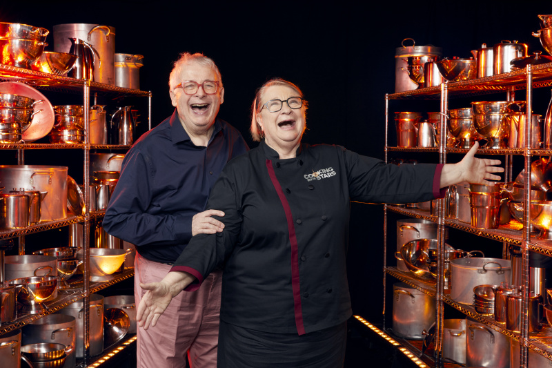  Ο Christopher Biggins και η μέντορά του Rosemary Shrager στέκονται ανάμεσα σε δύο ράφια εξοπλισμού μαγειρικής, και οι δύο χαμογελούν άγρια