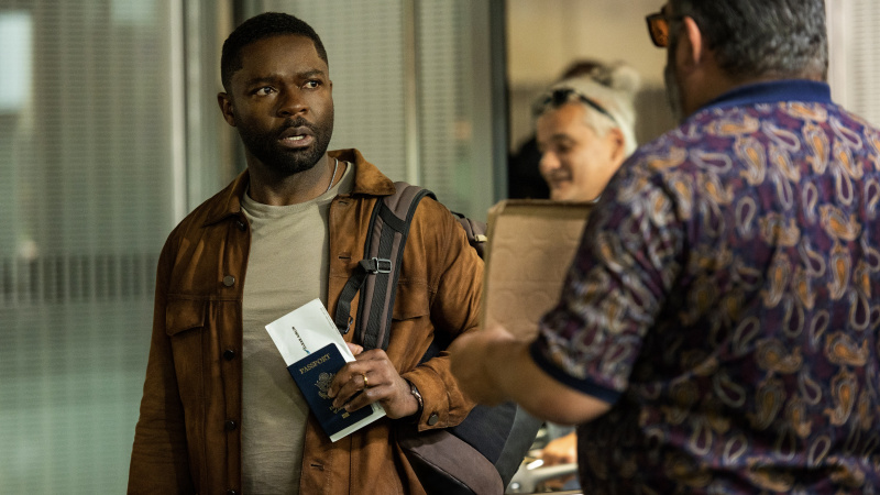   David Oyelowo, kahverengi ceketli, Dave'in pasaportu var ve Rol Oyunu'nda şaşırmış görünüyor.