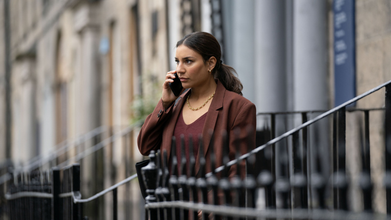   Lucie Shorthouse v hnedej bunde ako DC Siobhan Clarke telefonuje pri zábradlí v Rebuse.