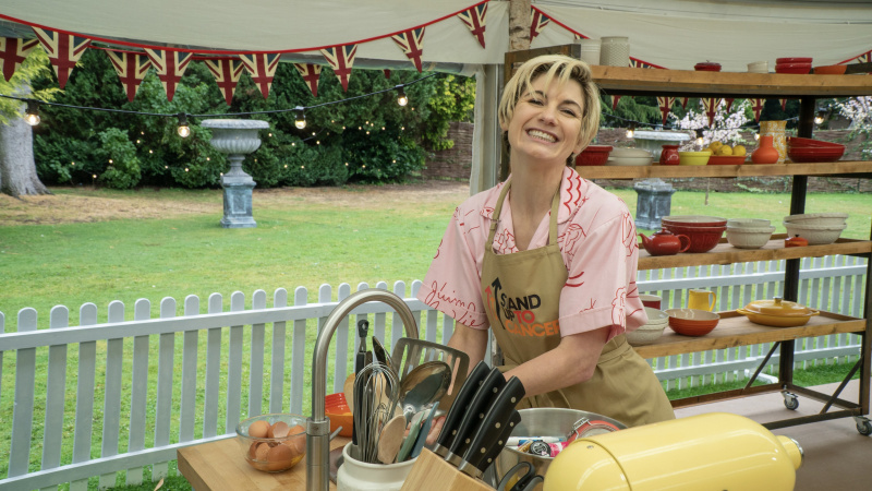   Jodie Whittaker v rožnati srajci in rjavem predpasniku Stand Up To Cancer stoji za svojo delovno mizo v The Great Celebrity Bake Off for Stand Up to Cancer