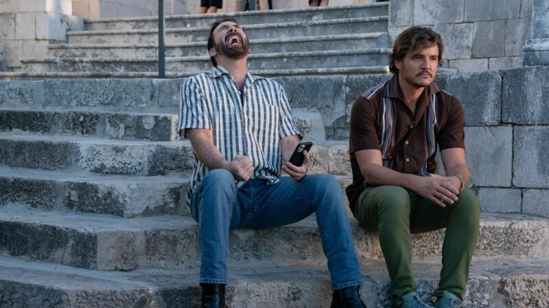   Nicolas Cage หัวเราะและนั่งถัดจาก Pedro Pascal ใน The Unbearable Weight of Massive Talent