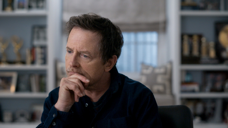  Uma cena de Still: A Michael J Fox Movie onde Michael J Fox está sendo entrevistado, de frente para a câmera. Ele está descansando o queixo na mão direita e olhando para o lado um pouco pensativo