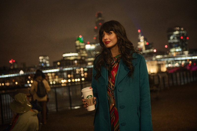   Jameela Jamil als verteller die een Starbucks-beker vasthoudt met de skyline van een stad achter haar.
