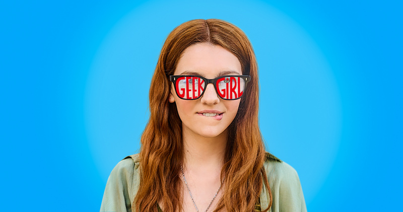 Harriet Carey puree huuliaan ja seisoo sinistä taustaa vasten nimilaseilla varusteltuna'Geek Girl' written across them in red.