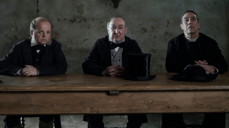   Dr. McBrearty (Toby Jones), Sir Otway (Dermot Crowley) e Padre Thaddeus (Ciaran Hinds) (da esquerda para a direita) sentados juntos em uma longa mesa em The Wonder