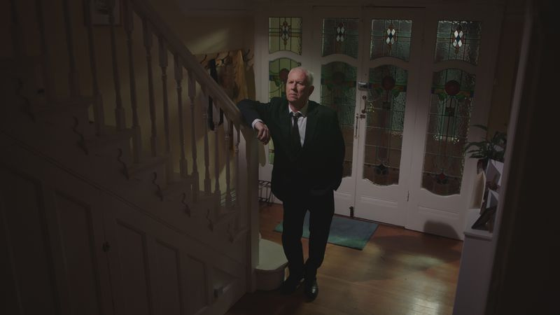 Charlie con un traje de funeral parado solo al pie de las escaleras en la casa que compartía con Duffy