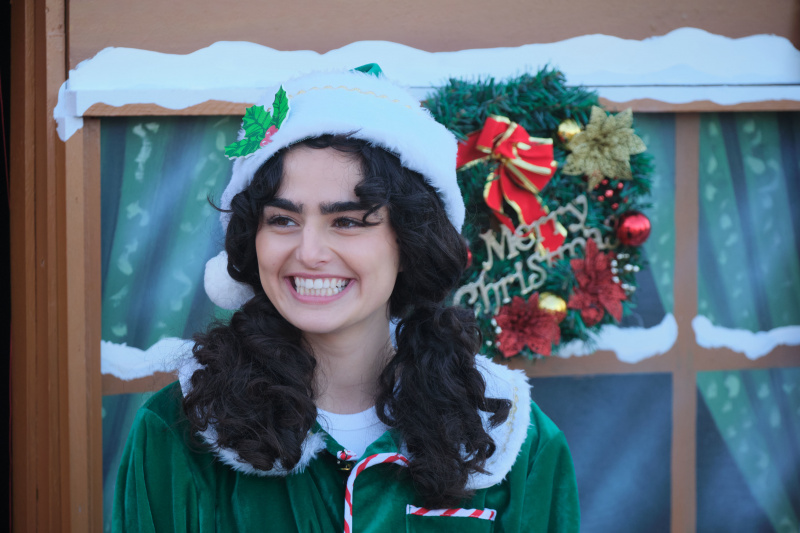   Zoe (Melina Sinadinou) steht vor dem Weihnachtsmann's grotto, dressed as an elf, smiling in a slightly forced way