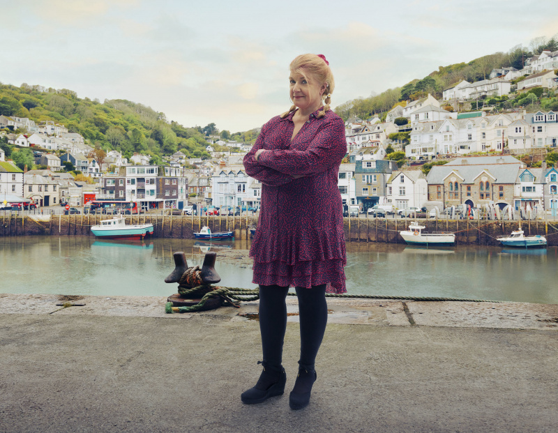   Eine Aufnahme von Felicity Montagu als Margo Martins in Beyond Paradise, wie sie im Hafen von Shipton Abbott steht, mit dem Meer und den Häusern, die im Hintergrund die Klippen hinaufragen