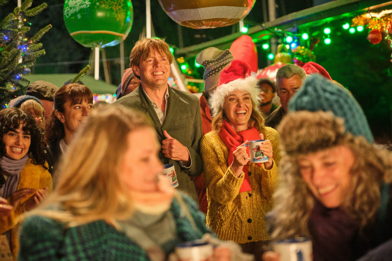   Ein Standbild aus dem Weihnachtsspecial „Beyond Paradise“, das Humphrey (Kris Marshall) und Martha (Sally Bretton) zeigt, wie sie in einer Menschenmenge auf dem Weihnachtsmarkt von Shipton Abbott stehen. Links von Humphrey in der Menge stehen Esther (Zahra Ahmadi) und ihre Tochter Zoe (Melina Sinadinou). Im hinteren Teil der Menschenmenge steht ein Weihnachtsbaum und darüber hängen große Dekorationen