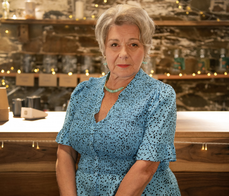   Barbara Flynn als Anne Lloyd in Beyond Paradise. Sie sitzt auf einem Hocker vor der Kamera bei Martha's café, facing the camera