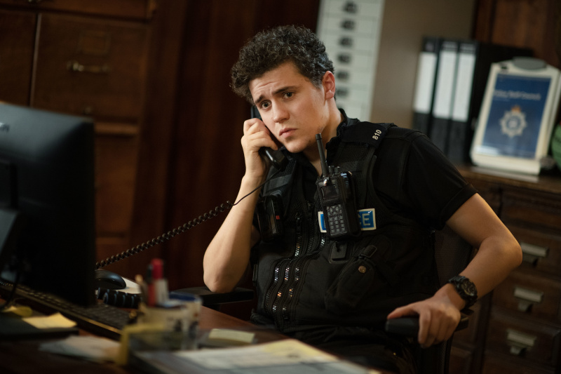   Dylan Llewellyn in der Rolle des PC Kelby Hartford. Er sitzt an seinem Schreibtisch auf der Polizeiwache und nimmt einen Anruf entgegen.