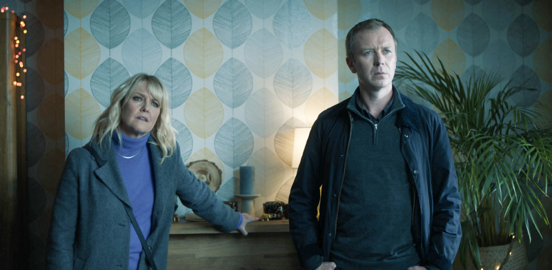   DI Ruth Calder (Ashley Jensen) och DC Sandy Wilson (Steven Robertson) förhör en misstänkt i sitt vardagsrum, medan Calder lutar sig mot mantelstycket
