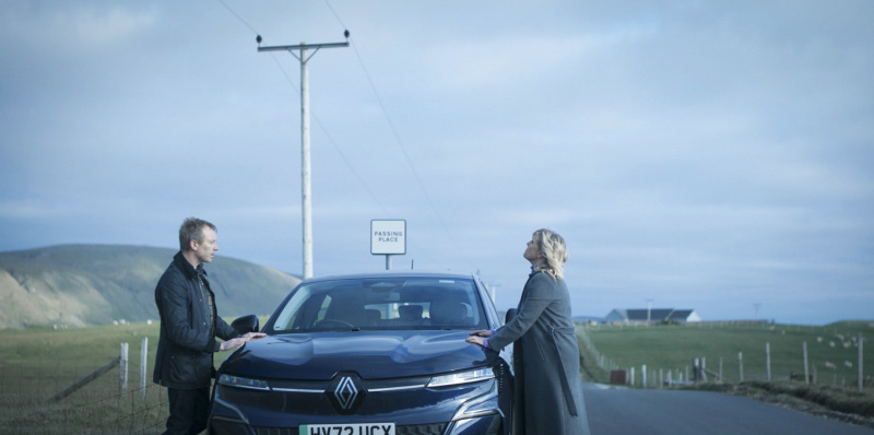   Sandy (Steven Robertson) och Calder (Ashley Jensen) står på varsin sida om en bil som står parkerad på en förbipasserande plats, båda lutade mot motorhuven. Calder ser upprörd och känslomässig ut.