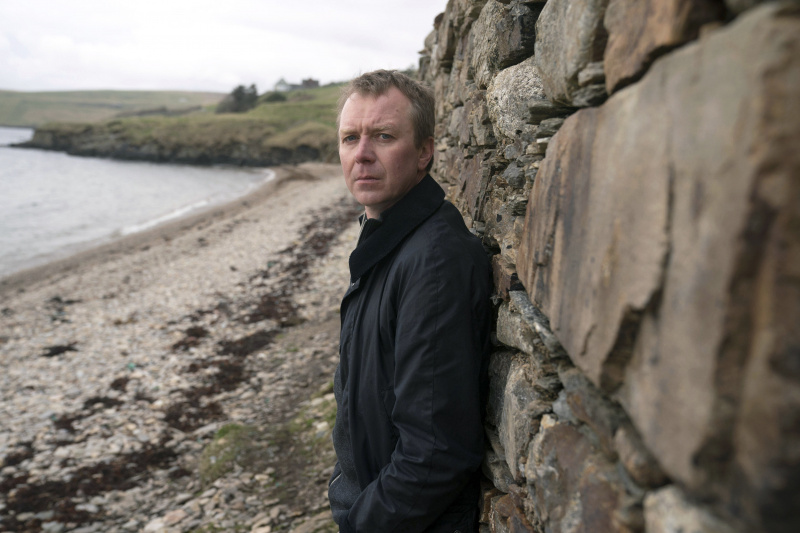   DC Sandy Wilson (Steven Robertson) står på en klapperstensstrand, lutad mot en tegelvägg bakom honom. Hans ansikte är vänt mot kameran.