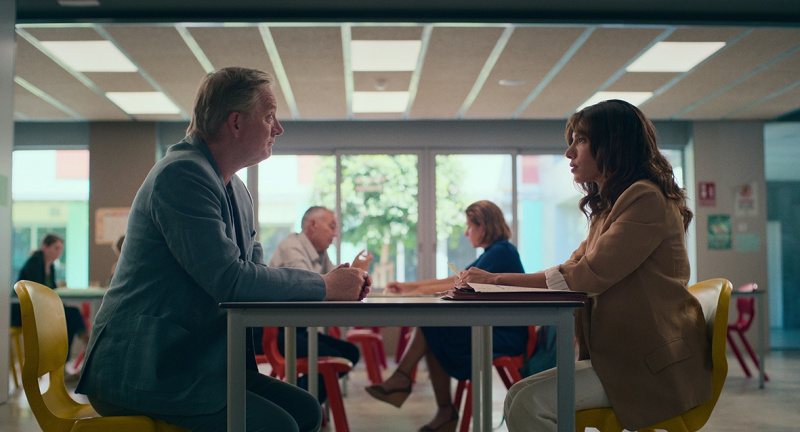   Daniel (Douglas Henshall) og Erin (Evin Ahmad) sitter på hver sin side av et bord på skolen og har et møte. Andre sammenkoblinger som har lignende møter er synlige bak dem.