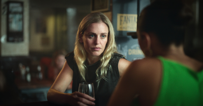  Lena (Denise Gough) sitter i en bar med et glass vin i hånden og snakker med en annen kvinne som har ryggen mot kameraet og er ute av fokus